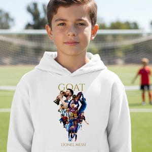 May include: White hoodie with a graphic of Lionel Messi, featuring the word "GOAT" in gold lettering. The design includes images of Messi in action. The hoodie is worn by a young person on a soccer field.