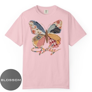 May include: A light pink short-sleeve t-shirt featuring a large butterfly graphic made of various patterned fabric patches. The butterfly has a brown textured body and frayed edges. Below the butterfly, the word Dolly is written in cursive script.
