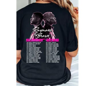 May include: A black t-shirt featuring a graphic of two men back-to-back with the text Raymond & Brown, Summer 2026, and a list of tour dates and locations printed in white and pink on the back of the garment, showcasing a concert tour design.