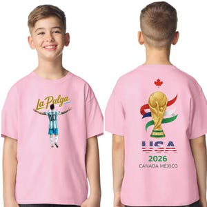 May include: Pink t-shirt featuring a soccer player graphic with the text "La Pulga" on the front. The back of the shirt displays a gold trophy, the USA flag, and the text "USA 2026 CANADA M&Eacute;XICO".
