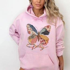 May include: A light pink hooded sweatshirt featuring a large butterfly graphic on the front. The butterfly wings are composed of various patchwork patterns in shades of blue, yellow, red, and white. The word Dolly is written in cursive below.