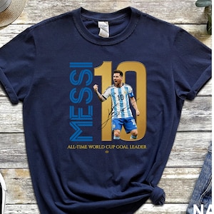 May include: A navy blue t-shirt featuring a graphic of Lionel Messi in an Argentina soccer jersey with the number 10. The design includes the text "MESSI", a large gold "10", a signature, and the text "ALL-TIME WORLD CUP GOAL LEADER" in gold.