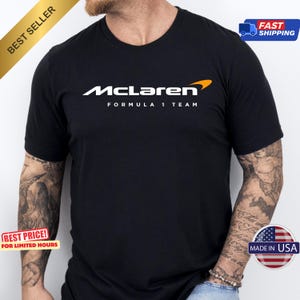 May include: Black t-shirt featuring the McLaren Formula 1 Team logo in white with an orange accent. The shirt has a crew neck and short sleeves. The text "BEST SELLER" is in the upper left corner. The shirt is made in the USA.