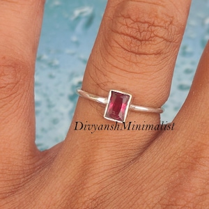 May include: A silver ring featuring a single rectangular, deep red gemstone set in a simple metal band. The ring is positioned on a finger, showcasing the clear, faceted cut of the stone and the smooth, polished finish of the silver setting.