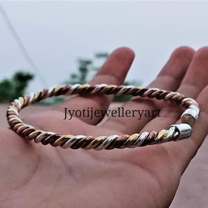 Op de afbeelding: Een gedraaide metalen armband met zilver-, goud- en kopertinten. De armband heeft een spiraalvormig ontwerp en een sluiting. De tekst "Jyotijewelleryart" is zichtbaar op de armband.