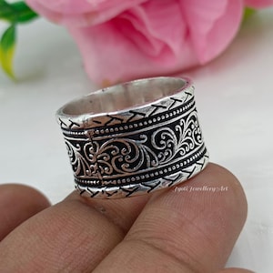 May include: A silver ring with an intricate, detailed design. The ring features a band with a repeating pattern of stylized floral and geometric elements. The ring is held by a person's fingers, with a pink flower in the background.