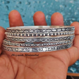May include: A set of four silver-toned metal bangles featuring distinct embossed patterns including geometric shapes, dots, and floral motifs. The bracelets are stacked together and display the text Divyansh Minimalist at the bottom.