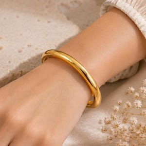 Pode incluir: Um bracelete rígido de tom dourado polido com uma superfície lisa e arredondada. A pulseira apresenta um design circular sólido e contínuo com um acabamento brilhante e reflexivo que capta a luz, criando uma estética limpa e simples.