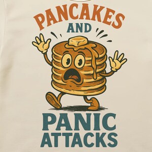 May include: Cream-colored t-shirt featuring a cartoon stack of pancakes with a surprised expression, butter on top, and the words "PANCAKES AND PANIC ATTACKS" in orange and teal. The design is a vintage style.