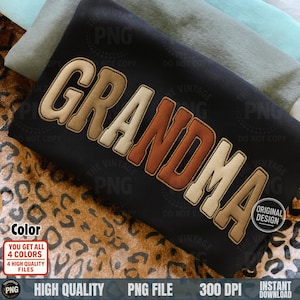 May include: A black t-shirt with the word "GRANDMA" in a layered, retro-style design. The text features a cream-colored outline, a tan middle, and a brown inner color. The shirt is folded on top of other folded shirts.