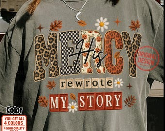 May include: A gray t-shirt with the words "MERCY His rewrote MY STORY" in a mix of leopard print, checkerboard, and pumpkin-themed lettering. The design includes fall leaves and daisy accents, with a "Original Design" stamp.