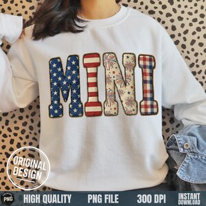 May include: White sweatshirt featuring the word "MINI" in a patriotic design. The letters are decorated with American flag patterns, fireworks, and red and blue plaid. The sweatshirt has a round neckline.
