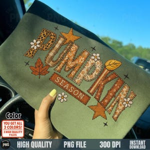 May include: A green sweatshirt with the words "PUMPKIN SEASON" embroidered in various patterns and colors, including floral, plaid, and leopard print. The design includes stars, leaves, and flowers, evoking an autumn theme.