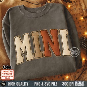 May include: A gray sweatshirt with the word "MINI" in large, textured letters across the front. The letters are in shades of beige and brown with a stitched outline. The design includes a "Original Design" badge.