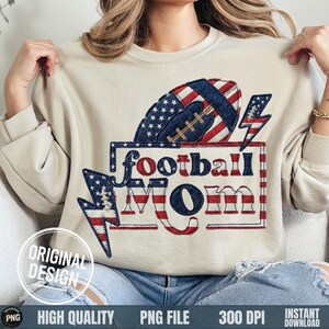 Puede incluir: Una sudadera beige con un diseño patriótico. El diseño presenta un balón de fútbol con un patrón de estrellas y rayas, las palabras "football Mom" en rojo, blanco y azul, y dos rayos con un patrón de estrellas y rayas. También se ven las palabras "Original Design".