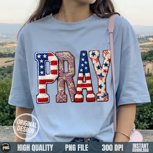 May include: Light blue t-shirt with the word "PRAY" in a patriotic design. The letters are decorated with red and white stripes, blue stars, and firework patterns. The shirt has short sleeves and a round neckline.
