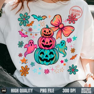 Może przedstawiać: Biała koszulka z kolorowym wzorem na Halloween. Wzór zawiera ułożone w stos dynie, duchy, nietoperze, kwiaty, gwiazdy i dużą kokardę. Napis "ORIGINAL DESIGN" znajduje się w okręgu po prawej stronie u góry.