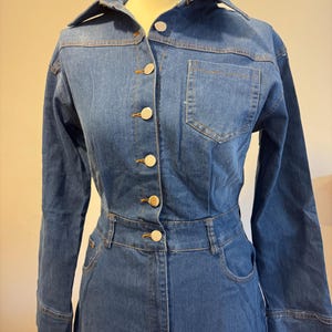 Op de afbeelding: Een blauwe spijkergoed jumpsuit met lange mouwen, een kraag en een knoopsluiting. De jumpsuit heeft een getailleerde taille, een borstzak en goudkleurige knopen. Het kledingstuk wordt op een mannequin gepresenteerd.