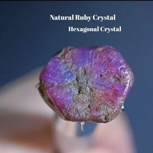 May include: A natural ruby crystal with a hexagonal shape and a deep magenta and purple hue. The surface features a rough, textured appearance with visible mineral inclusions. The text Natural Ruby Crystal Hexagonal Crystal is visible.