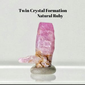 May include: A natural ruby twin crystal formation with a pink, translucent, and textured surface. The crystal features a vertical main body with a smaller horizontal protrusion, resting on a smooth, rounded grey stone base. Text reads: Twin Crystal Formation Natural Ruby.