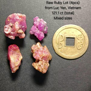 Op de afbeelding: Vier ruwe robijn edelstenen uit Luc Yen, Vietnam, met een totaal gewicht van 121,1 karaat. De robijnen zijn in verschillende tinten roze en rood, met een goudkleurige munt in de buurt. De tekst op de afbeelding luidt "Raw Ruby Lot (4pcs) from Luc Yen, Vietnam 121.1 ct (total) Mixed sizes."