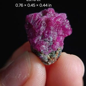 Peut inclure: Un amas de rubis brut de Luc Yen, Vietnam, d'un rose profond. La gemme mesure 1,93 x 1,14 x 1,12 cm et pèse 23,10 ct. La surface rugueuse et texturée du rubis est visible sur un fond sombre.