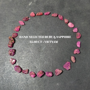 May include: A collection of small, raw, pinkish-red ruby and sapphire gemstones arranged in a circle. The text in the center reads: HAND-SELECTED RUBY & SAPPHIRE 35.60 CT • VIETNAM. These natural, unpolished stones vary in shape and size.