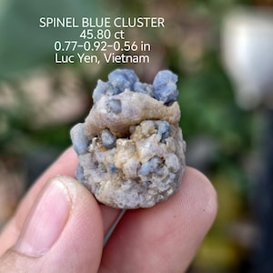 Może przedstawiać: Ręka trzyma niebieski klaster spinelu. Tekst na zdjęciu brzmi: SPINEL BLUE CLUSTER, 45,80 ct, 1,96-2,34-1,42 cm, Luc Yen, Wietnam. Klaster to mieszanka jasnoniebieskich i beżowych kolorów.