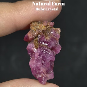 May include: A raw, natural form ruby crystal with a deep magenta-pink hue and irregular, faceted surfaces. The stone features textured, brownish-tan mineral inclusions at the top. The text 'Natural Form Ruby Crystal' is printed in white above the stone.