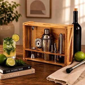 Op de afbeelding: Een houten bar-organizer met een roestvrijstalen cocktailshaker, een jigger, een zeef, een stamper en een barlepel. De set wordt getoond met een glas mojito, twee boeken met de titels The Home Bar en Cocktails, en een wijnfles.