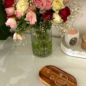 Op de afbeelding: Een boeket rozen in rood, roze en geel in een heldere glazen vaas. Een houten ovaal dienblad bevat sieraden, waaronder ringen en een armband. Een witte kandelaar en een kleine kaars zijn ook zichtbaar.