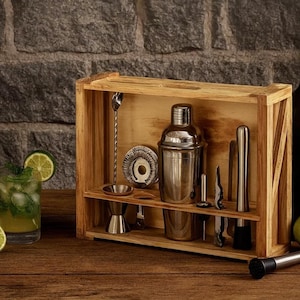 Pode incluir: Um conjunto de ferramentas de bar de madeira com uma coqueteleira, um pilão e outras ferramentas de aço inoxidável. Uma garrafa de vidro escuro com uma rolha, um coquetel e limas também estão presentes. O conjunto é exibido em uma superfície de madeira.