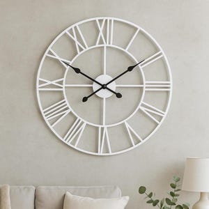 May include: A large, white, open-faced wall clock with black hands and Roman numerals. The clock has a circular design with a minimalist aesthetic. The clock is mounted on a light gray wall.