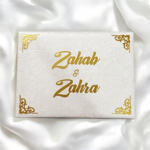 May include: A rectangular, off-white box with gold lettering that reads "Zahab & Zahra." The box has gold decorative corner accents. The box is resting on a white, silky fabric.