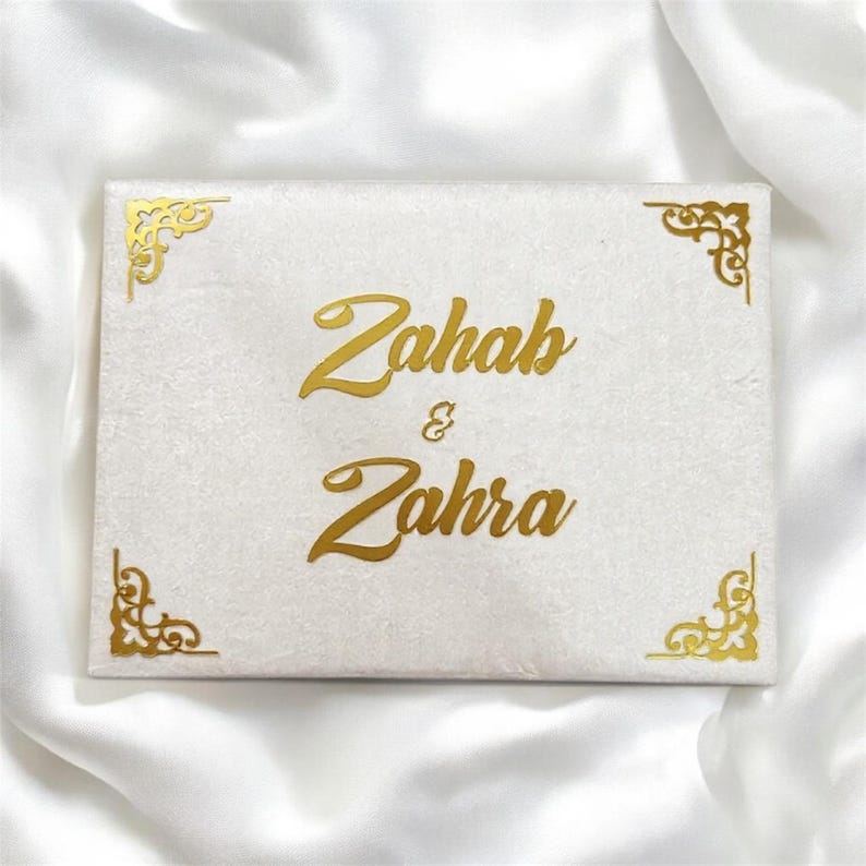 May include: A rectangular, off-white box with gold lettering that reads "Zahab & Zahra." The box has gold decorative corner accents. The box is resting on a white, silky fabric.