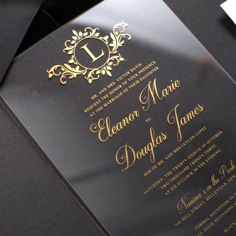 Pode incluir: Um convite de casamento em acr&iacute;lico transparente com letras em folha de ouro e um bras&atilde;o decorativo com monograma com a letra L. O texto inclui os nomes Eleanor Marie e Douglas James, juntamente com detalhes do evento, data e local.