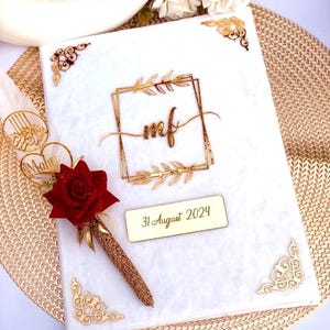 May include: A white wedding guest book with gold detailing, displaying the initials "mf" inside a gold frame. A pen with a red rose and the date "31 August 2024" are included. Decorative gold corner pieces and a gold feather enhance the elegant design.