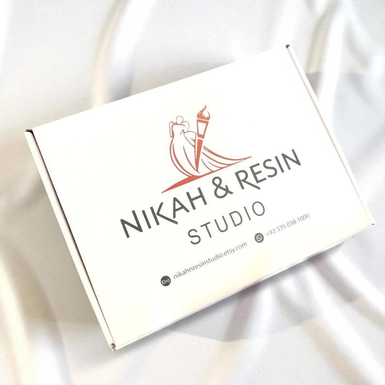 May include: A white cardboard box with the text "NIKAH & RESIN STUDIO" in dark gray. The logo features a stylized image of a bride and a torch in orange. The Etsy shop's URL and phone number are also visible.