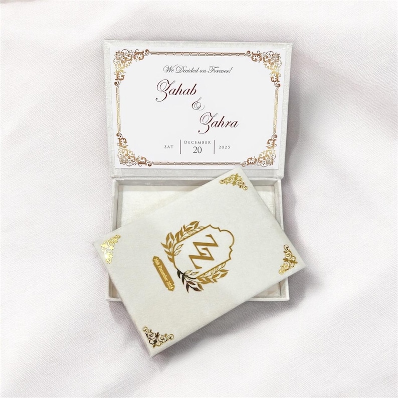 May include: A white wedding invitation box with gold accents. The box is open, revealing a card with the names "Zahab & Zahra" and the date "December 20, 2023". The card inside features the initials "ZZ" in gold.