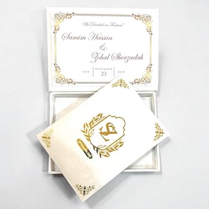 May include: A white wedding invitation box with gold decorative accents and text. The invitation card features the names "Samim Hasan & Zohal Sherzadah" and the date "November 23, 2025". The card inside has a gold emblem.