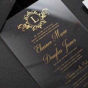 Pode incluir: Um convite de casamento em acr&iacute;lico transparente com letras em folha de ouro e um monograma decorativo com a letra L. O texto detalha o casamento de Eleanor Marie e Douglas James no s&aacute;bado, vinte e oito de outubro de dois mil e vinte e tr&ecirc;s.