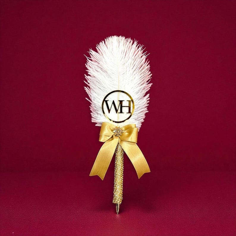A decorative feather pen featuring a white feather plume, a gold bow, and a gold initials of bride and groom. It is used for to sign the Nikkah certificate and also called Nikkah Pen, Qubool Hai pen.