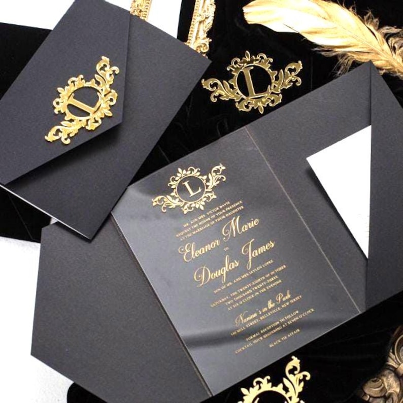 Pode incluir: Um conjunto de convites de casamento preto com um cart&atilde;o de acr&iacute;lico transparente com letras em folha de ouro e um envelope preto a condizer. O envelope e o cart&atilde;o s&atilde;o adornados com um emblema de filigrana dourada com a letra L.