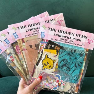 May include: A hand holding several clear plastic bags filled with ephemera. Each bag has a pink and white label that reads "THE HIDDEN GEMS EPHEMERA PACK Y2K EDITION 25 UNDISCOVERED ODDS & ENDS". The bags contain stickers, fabric, and other decorative items.