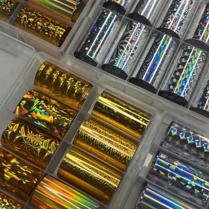 Op de afbeelding: Een verzameling nail art folies in diverse kleuren en patronen, waaronder goud, zilver en holografische ontwerpen. De folies zijn opgerold en opgeslagen in doorzichtige plastic containers, met een reeks texturen en effecten voor nail art.
