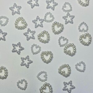 May include: Scattered white pearl embellishments in star and heart shapes. The pearls are arranged to form the outlines of the shapes, creating a delicate and elegant appearance. These decorative elements are ideal for craft projects.