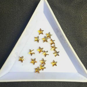 May include: A collection of small, gold-colored star-shaped embellishments inside a white triangular tray. The stars are scattered, reflecting light. The tray sits on a dark, textured surface. These are likely decorative craft supplies.