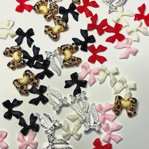 May include: An assortment of decorative nail art charms in various designs. The charms include small bows in red, black, pink, and white, along with leopard print bows and silver bow-shaped charms with rhinestones. The charms are scattered on a white surface.