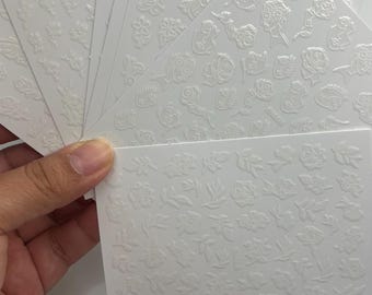 Puede incluir: Una mano sostiene varias hojas de pegatinas blancas para decoración de uñas. Cada hoja presenta diseños florales y de hojas en relieve. Las pegatinas tienen varias formas y tamaños, incluyendo rosas, hojas y otros elementos botánicos. Las pegatinas están sobre un fondo blanco.