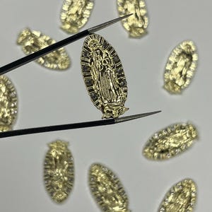May include: Gold-colored oval-shaped charms featuring a religious figure, held by tweezers. The charms are detailed with intricate designs and small embellishments. Several other charms are scattered around on a white surface.